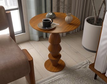 May include: A small, round wooden side table with a unique stacked base. The tabletop holds a black coffee cup on a saucer, a small white vase, and a decorative lamp. The table is approximately 24 inches tall.