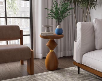 May include: A wooden side table with a round top and a geometric base. The table is brown with a yellow accent. A vase with blue and white accents and greenery sits on top, along with a small gray dish and two books. A brown chair and white sofa are in the background.