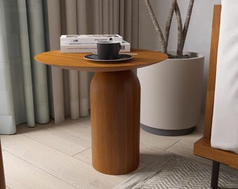 May include: A round, wooden side table with a dark brown finish. A black coffee cup and saucer sit on top, along with two books. The table has a cylindrical base and is placed near a potted plant and a bed frame.