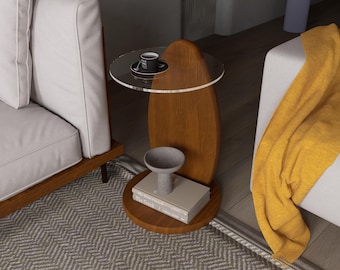 May include: A modern side table with a clear circular top and a wooden base. A small black cup and saucer sit on the top. The table has a wooden oval-shaped center and a round base. A small gray bowl and books are on the lower shelf.
