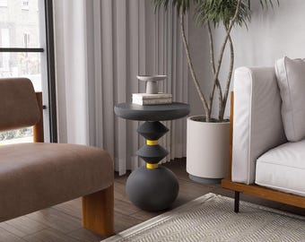 May include: A modern, dark gray side table with a unique geometric design. The table features a round top, stacked conical shapes, and a spherical base, accented with yellow bands. A small plant and books rest on top, with a neutral rug and sofa in the background.