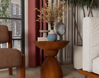 May include: A wooden side table with a unique hourglass-shaped base. The table is topped with a vase of dried flowers, a blue and white vase, a gray Buddha head statue, and a small stone sculpture. The table is in a living room setting.