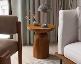 May include: A wooden side table with a round top and a fluted base. The table is made of light brown wood and has a small lamp, a vase with dried flowers, and a decorative stone on top. The table is in a living room setting.