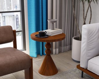 May include: A small, round wooden side table with a conical base and a stacked ball design. A black coffee cup and saucer sit on top, along with books and a decorative bowl. The table is near a window with blue curtains.