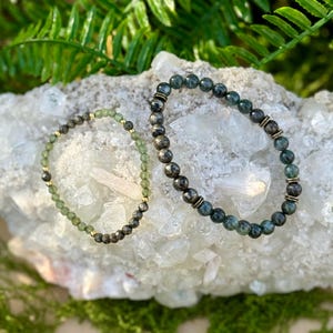 May include: Two beaded bracelets with green and dark gray stones. One bracelet has gold-colored accents. The bracelets are displayed on a white crystal formation, surrounded by green ferns and moss.