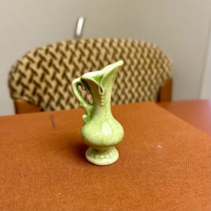 May include: A miniature green ceramic jug with a handle and a flared rim. The jug has a rounded body and a pedestal base. The colour is a light green with darker green detailing. It is likely a decorative item.