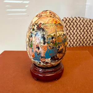 May include: An egg-shaped decorative item with a detailed, colorful scene of people in traditional clothing. The item is mounted on a dark red wooden stand. The artwork features a variety of colors, including blue, orange, and green.
