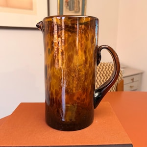 May include: A tall, amber-colored glass pitcher with a handle and a spout. The pitcher has a speckled pattern throughout the glass. The handle is a dark brown color. The pitcher is sitting on a reddish-orange surface.