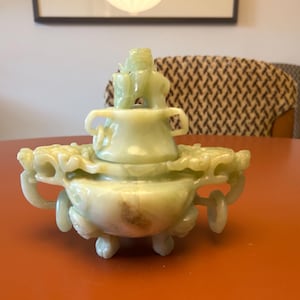 Puede incluir: Un quemador de incienso de piedra tallada de color verde pálido con intrincados detalles florales y de dragón. La pieza decorativa tiene una tapa y pequeñas asas, que descansa sobre una base redonda con pequeños pies. El quemador se exhibe sobre una mesa naranja.