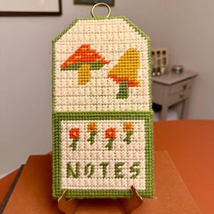 May include: A handmade, off-white needlepoint notepad holder with a green border. The top section features two mushroom designs in orange, yellow, and green. The bottom section has the word "NOTES" and floral designs. A gold-colored ring is at the top.