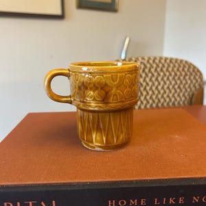 May include: A small, brown ceramic coffee mug with a handle. The mug has a textured design around the top and a geometric pattern on the lower portion. It sits on a stack of books with the word "CAPITAL" visible.