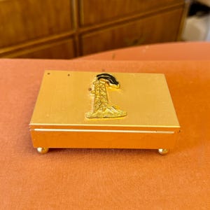 May include: A small, rectangular gold-colored box with a decorative oil derrick emblem on the lid. The box has rounded feet and a hinged lid. The oil derrick emblem is gold with black accents.