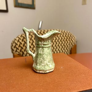 May include: A small, antique-style ceramic jug with a handle. The jug is primarily green with white speckles and a ruffled rim. The base is a light beige colour. The jug is sitting on a brown surface.