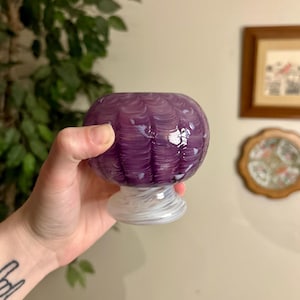 Puede incluir: Un cuenco de vidrio morado con un diseño de remolino blanco y morado. El cuenco tiene forma redondeada y se asienta sobre una base blanca. El vidrio tiene un acabado brillante.