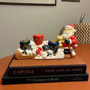 Puede incluir: Un portavelas navideño festivo con Papá Noel y tres candeleros. Papá Noel viste un traje y un gorro rojos. Los candeleros son rojos, verdes y azules. La base es un tronco con nieve y acebo.