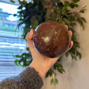 Könnte beinhalten: Ein poliertes, kugelförmiges Dekorationsobjekt mit einem satten, rotbraunen Farbton und aufwendigen goldenen Adern. Die Kugel wird in einer Hand gehalten, wodurch ihre glatte, reflektierende Oberfläche zur Geltung kommt. Das Design der Kugel deutet auf ein dekoratives Element für ein Zuhause oder Büro hin.