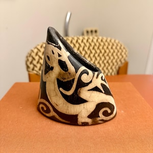 May include: A black and cream-colored, triangular-shaped decorative object. The object features a stylized design of a creature with swirling patterns. The object is likely made of a hard material, possibly ceramic or stone.