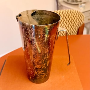 May include: A vintage silver-coloured jug with a handle and a curved spout. The jug has a cylindrical body and a tarnished, textured surface. The handle is a simple, angular design. The jug is sitting on a brown surface.