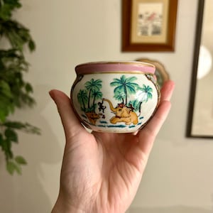 Peut inclure: Un petit bol rond en céramique avec un bord rose et une illustration détaillée de palmiers, d'un éléphant et d'un singe. Le bol est blanc avec une bordure dorée.