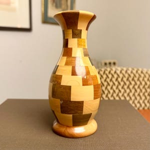 May include: A decorative wooden vase with a mosaic pattern of light and dark brown squares. The vase has a wide base, a rounded body, and a flared top. The vase is approximately 10 inches tall.