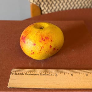 Op de afbeelding: Een gele appel met rode vlekken, liggend op een bruin oppervlak. Een houten liniaal met inch-markeringen staat op de voorgrond. De appel heeft een steel en een enigszins onregelmatige vorm.