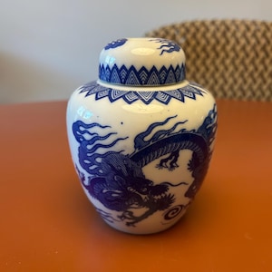 Puede incluir: Un tarro de jengibre de cerámica blanca y azul con tapa. El tarro presenta un diseño detallado de dragón azul y patrones decorativos. El tarro está sobre una superficie naranja.