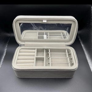 Puede incluir: Estuche de viaje para joyas gris claro abierto, con un espejo dentro de la tapa. El estuche tiene múltiples compartimentos para anillos, aretes y pulseras. La cremallera es plateada. La marca "PANDORA" es visible en el espejo.