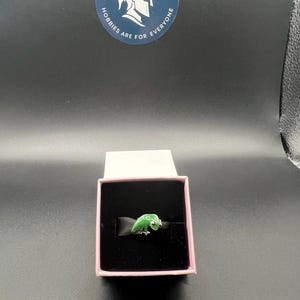 May include: A green enamel ring in the shape of a lizard, presented in a pink jewelry box with a black interior. The ring is set against a black ribbon. The box is open, revealing the ring.