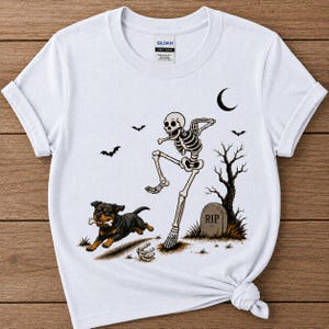 May include: A white short-sleeve t-shirt featuring a graphic print of a skeleton dancing near a tombstone marked RIP, a bare tree, bats, a crescent moon, and a black and tan Rottweiler dog running with a bone in its mouth.