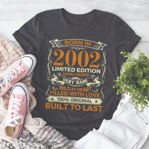 May include: Dark gray t-shirt with orange text that reads "BORN IN 2002 LIMITED EDITION LIVING LEGEND VERY RARE WILD AT HEART FILLED WITH LOVE 100% ORIGINAL BUILT TO LAST." Pink canvas sneakers are on the left.