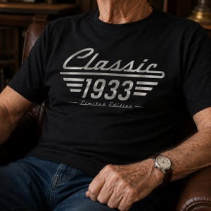 Puede incluir: Camiseta negra con la inscripción plateada "Classic 1933 - Limited Edition". La camiseta de manga corta parece estar hecha de un material suave. La persona también lleva un reloj con una correa de cuero marrón.