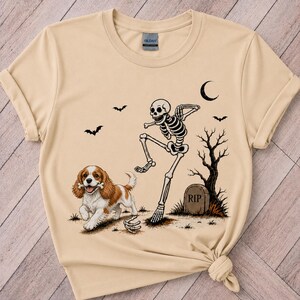 以下が含まれることがあります： サンドカラーのTシャツ。口に骨をくわえた茶色と白のキャバリア・キング・チャールズ・スパニエル、踊る骸骨、RIPと刻まれた墓石、枯れ木、三日月、飛んでいるコウモリのグラフィックプリントが施されています。