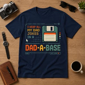 Könnte beinhalten: Marineblaues T-Shirt mit einem pixeligen Design, das eine Diskette und den Text "I KEEP ALL MY DAD JOKES IN A DAD-A-BASE" zeigt. Das Design enthält ein Raster, einen Cursor und die Worte "100% SAVED".