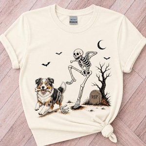 Peut inclure: Un t-shirt couleur crème avec une impression graphique représentant un chien berger australien tricolore tenant un os dans sa gueule, accompagné d'un squelette dansant, d'une pierre tombale marquée RIP, d'un arbre nu, d'un croissant de lune et de chauves-souris.