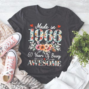 Könnte beinhalten: Ein dunkelgraues Kurzarm-T-Shirt mit dem Text Made in 1966 60 Years Of Being AWESOME. Die Zahlen und das Wort AWESOME sind mit einem bunten Blumenmuster gefüllt. Kleine rote Herzen und ein Blumenarrangement verzieren das Design.