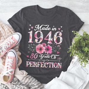 Op de afbeelding: Donkergrijs T-shirt met de tekst "Made in 1946" en "80 Years Of PERFECTION" in wit en bloemmotieven. Roze sneakers en een witte jeans zijn zichtbaar. Een kleine groene plant staat ook in de afbeelding.