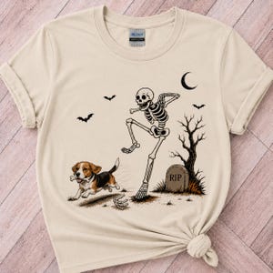Może przedstawiać: Beżowy t-shirt z okrągłym dekoltem z nadrukiem przedstawiającym tańczący szkielet obok nagrobka z napisem RIP i bezlistnego drzewa, z biegnącym psem rasy beagle trzymającym w pysku kość, w towarzystwie nietoperzy i półksiężyca na niebie.