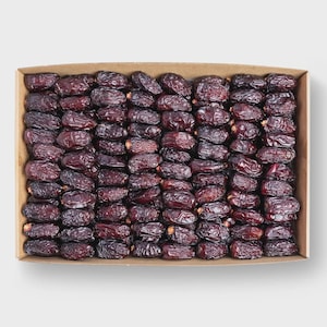 May include: A rectangular cardboard box filled with a dense, organised arrangement of dark brown, wrinkled dried dates. The fruit is packed tightly in rows, showcasing a uniform, deep mahogany colour and a textured, shrivelled skin surface.