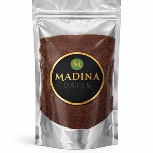 May include: A silver pouch filled with brown granules, likely a food product. The pouch has a circular label with "MADINA DATES" in white text, a gold border, and a green "M" logo. The packaging is sealed, suggesting freshness and preservation of the contents.