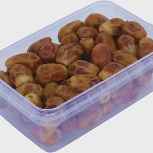 May include: A rectangular, clear plastic container filled with numerous whole, dried brown dates. The dates have a wrinkled, textured skin with varying shades of light and dark brown, and they are packed together inside the transparent storage box.