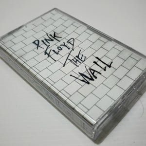 May include: A clear cassette tape case with a white and gray brick wall design. The text "PINK FLOYD THE WALL" is handwritten in black marker across the front of the case. The cassette tape is a vintage music collectible.