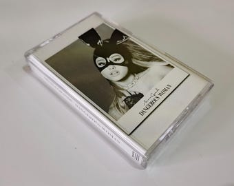 May include: A cassette tape featuring the album cover for "Dangerous Woman" by Ariana Grande. The cover art is a black and white image of a woman wearing a bunny mask. The album title and artist name are printed on the cover.