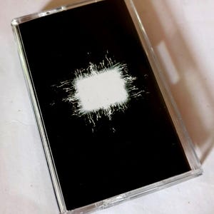 May include: A black cassette tape with a white rectangular design in the center, surrounded by a white, textured pattern. The cassette is encased in a clear plastic case.