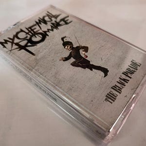 Könnte beinhalten: Eine Musikkassette für das Album "The Black Parade" von My Chemical Romance. Das Cover zeigt das Logo der Band und eine Cartoonfigur in schwarzer Uniform. Die Kassette befindet sich in einer transparenten Kunststoffhülle.
