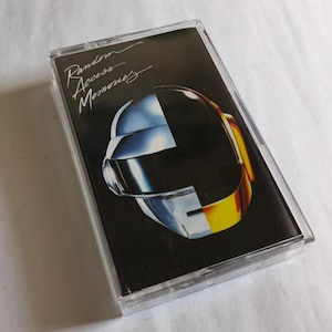 May include: A cassette tape of the album "Random Access Memories" by Daft Punk. The cover features a black background with a stylized helmet design in silver, black, and gold. The album title is written in white script.