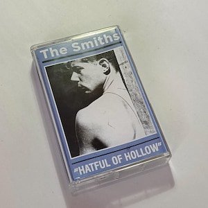 May include: A cassette tape for "Hatful of Hollow" by The Smiths. The clear cassette case has a blue and white label with a black and white image of a man. The text "The Smiths" and "Hatful of Hollow" are printed on the label.