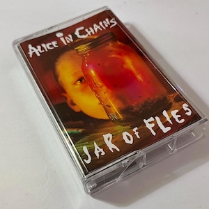 Könnte beinhalten: Eine Musikkassette mit dem Albumcover von Alice in Chains' "Jar of Flies". Das Cover zeigt ein Gesicht, das teilweise von einem Glas mit einer rötlichen Flüssigkeit verdeckt wird. Der Albumtitel ist in weißer Schrift.