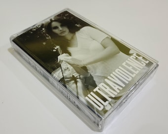 Puede incluir: Un casete en una funda de plástico transparente. La portada presenta una imagen en tonos sepia de una persona y la palabra "ULTRAVIOLENCE" en blanco. El casete está sellado en plástico.