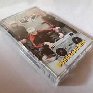 May include: A cassette tape of the Beastie Boys' "Solid Gold Hits" album. The cover features a photo of the band members with a boombox. The text "Solid Gold Hits" is in yellow, and the band's name is in a diamond-shaped logo.