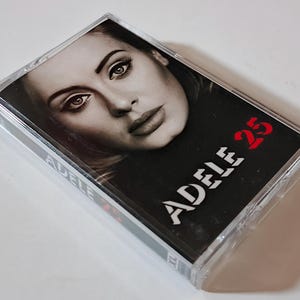 May include: A clear plastic cassette tape case featuring the album cover for Adele 25. The cover art displays a black and white portrait of a person with the text ADELE 25 printed in bold red letters on the lower right side of the insert.